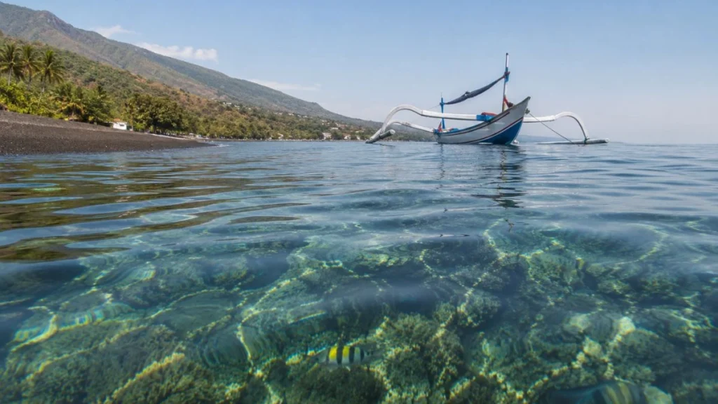 Amed Bali vs Candidasa snorkeling — eye-level view of clear blue-green water at black sand beach with coral visible below surface and jukung boat offshore.