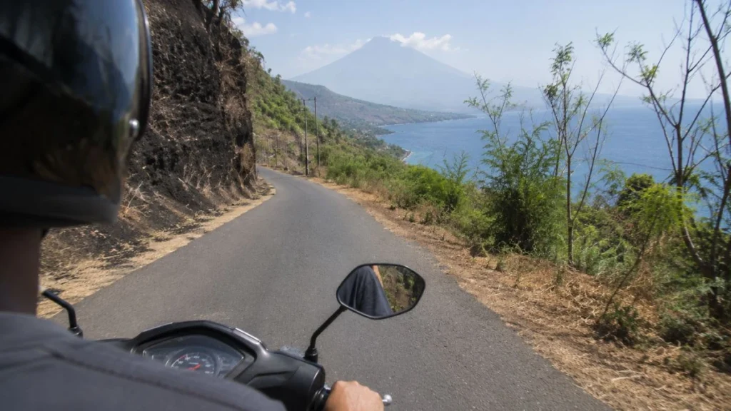 Bali road trip east bali — over-the-shoulder scooter view on narrow coastal road with ocean to the right and Mount Agung outline ahead in midday haze.