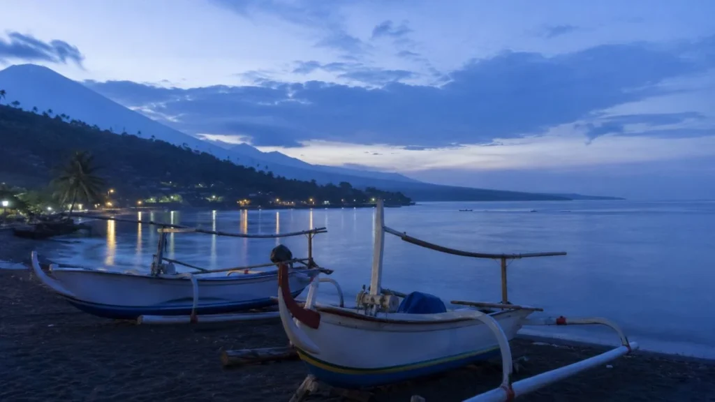 Best beaches in Bali for slow travellers — Jemeluk bay Amed at pre-dawn with jukung fishing boats on black volcanic sand and calm bay reflecting soft blue sky.